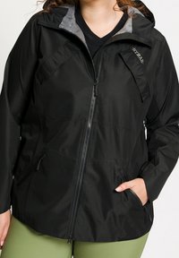 Kvinna i en svart huva jacka med dragkedja och fickor, handen i fickan, över en svart skjorta och gröna byxor.