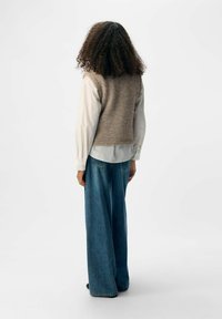 Brauner Strickwesten über einem weißen Hemd mit Knöpfen, kombiniert mit weit geschnittenen blauen Jeans. Schlichtes Design mit strukturierter Oberfläche.