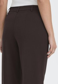 Pantalons marron taillés avec une texture lisse, dotés d'une taille ajustée et de poches latérales. Le tissu est uni, sans motifs ni ornements.