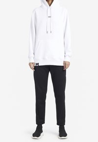 Persona che indossa una felpa bianca con cappuccio e tasca frontale, pantaloni neri a sigaretta e sneakers nere senza lacci con suole bianche, in piedi.