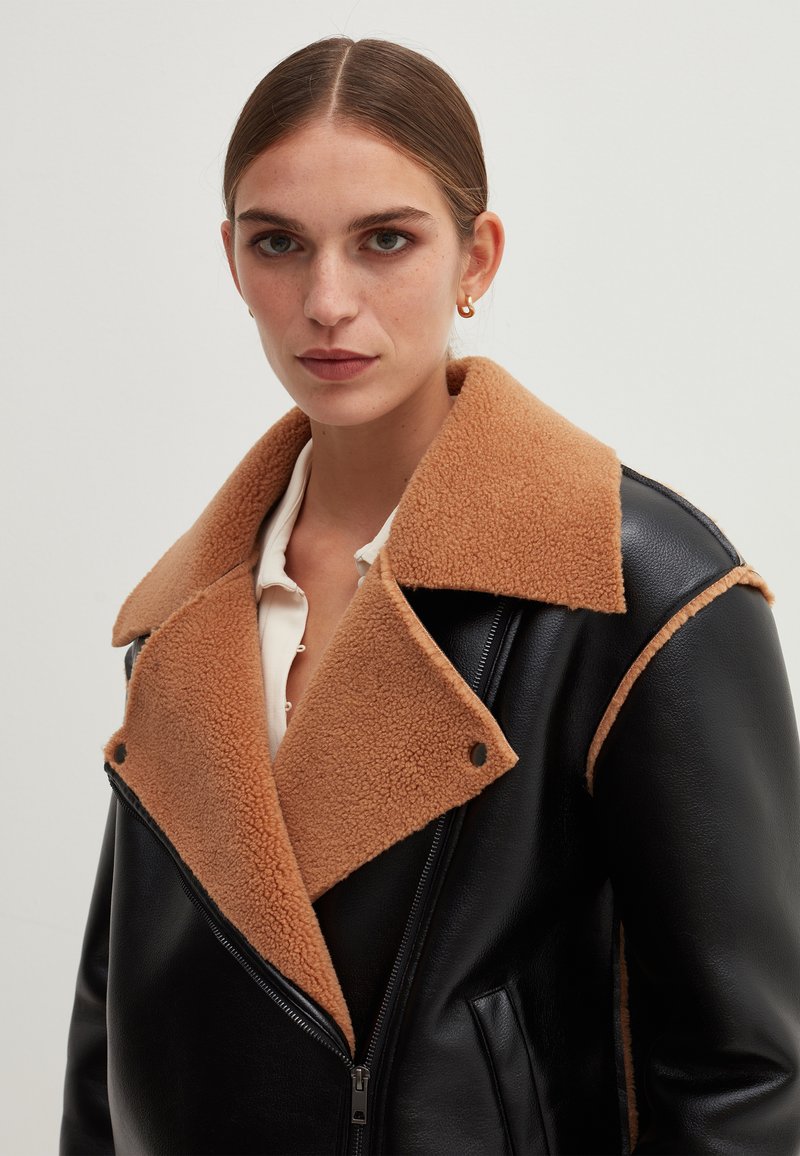 Schwarze Lederjacke mit einem tanfarbenen Schafslederkragen, ausgestattet mit einem doppelt strukturierten Design, Reißverschluss und seitlichen Taschen.
