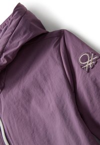 Giacca viola con cappuccio e patch logo circolare ricamato sulla manica superiore, mostrata su uno sfondo bianco.