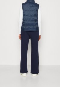 Granatowa kamizelka puchowa z wysokim kołnierzem i pikowanym wzorem, zestawiona z granatowymi spodniami typu wide-leg i białym obuwiem, widziana z tyłu.
