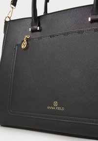 Black textured tote bag with gold hardware, featuring a zippered pocket, brand logo in gold, and dual top handles.
