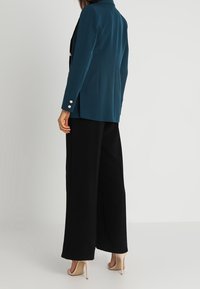 Morski zielony blezer o dopasowanym kroju, z wyciętym kołnierzem, ozdobionymi guzikami na rękawach, zestawiony z czarnymi, szerokimi spodniami.