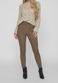 Kvinna klädd i en beige stickad tröja, bruna rutig slim-fit byxor och svarta ankelboots som står mot en grå bakgrund.