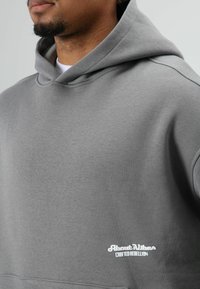 Grijze capuchontrui met een gladde textuur, voorzien van een wit geborduurd logo linksonder. Eenvoudig ontwerp, zachte stof en een casual pasvorm.