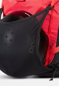 Tasca in rete nera di uno zaino con un casco nero arrotondato. Lo zaino è realizzato in tessuto rosso testurizzato con motivi diagonali e spallacci regolabili.