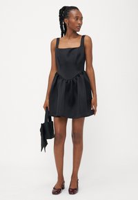 Melna satīna kleita ar kvadrātveida dekoltē un platām lencēm, kurai ir savilkts svārks. Valkāta ar mazu melnu rokassomu un bordo kurpēm.