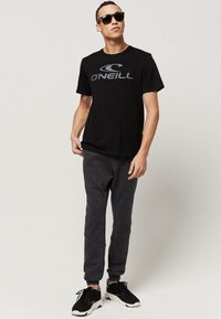 Sort O'Neill t-shirt med grå logo, grå joggingbukser og sorte sneakers med hvide såler. Model med solbriller, står afslappet.