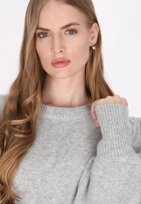 Szary, dziany sweter z szerokim okrągłym dekoldem i ściągaczami przy rękawach. Miękka struktura i luźny krój. Włosy modelki są falowane i opadają na jedno ramię.