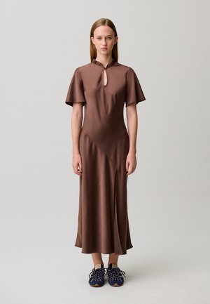 Donna in piedi di fronte, indossa un vestito midi marrone con maniche svolazzanti e sneakers blu navy su uno sfondo semplice grigio chiaro.