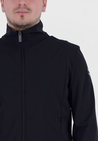Giacca nera con zip alta, texture liscia e tasca laterale con zip. Presenta un logo sulla manica sinistra.