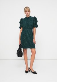Teal textured dress with puff sleeves, adorned with sequins and fringe at the hem. Paired with a black bag and pointed black heels.