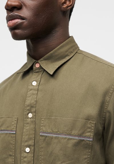 Πουκάμισο σε ελαιώδες πράσινο χρώμα με γιακά, δύο τσέπες στο στήθος και διακοσμητική πολύχρωμη ρίγα στα άκρα των τσεπών. Διαθέτει κρεμ κουμπιά.