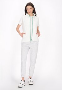 Canotta bianca senza maniche con zip verde e tasche, abbinata a pantaloni felpa grigi chiari e sneakers bianco e nere. Design semplice e sportivo.