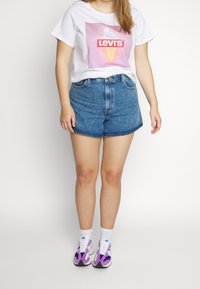 Bílý tričko s růžovou grafikou a červeným logem "Levi's", doplněné modrými džínovými šortkami. Nosené s fialovými a bílými teniskami.