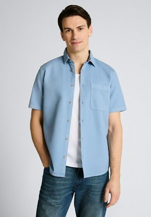Mężczyzna w jasnoniebieskiej, krótkiej koszuli z guzikami na białym podkoszulku, w niebieskich dżinsach, stojący na jednolitym jasnoszarym tle.