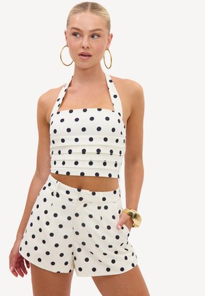 Femme portant un crop top blanc à col halter et un short à pois noirs, boucles d'oreilles anneaux dorés et un large bracelet en or, posant avec la main dans la poche.