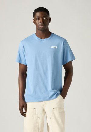 Mężczyzna ubrany w jasnoniebieski t-shirt Levi's i kremowe spodnie, stojący z jedną ręką w kieszeni na jednolitym tle.