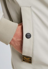 Beige jacket featuring textured fabric, ribbed cuffs, a patch pocket with black button, and a branded label in gold and black.