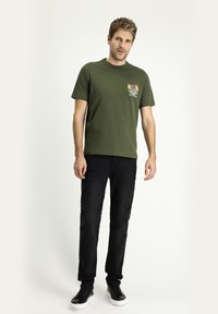 Camiseta de algodón verde oliva con cuello redondo y mangas cortas, con un diseño gráfico en el lado izquierdo del pecho. Combinada con jeans negros y zapatillas negras.