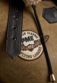 Bruin leren patch met zwarte band, voorzien van geborduurd logo met vleugels en "Flying Legend." Gelaagde achtergrond met stiksel details.