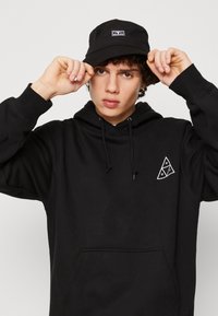 Sort hættetrøje med snore, frontlomme og hvid geometrisk logo. Kombineret med en sort bøttehat med et lille logo patch.