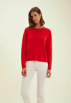 Pull rouge à manches longues en matière douce, avec un col ras du cou et un petit logo "IF" près de l'ourlet. Associé à un pantalon blanc.