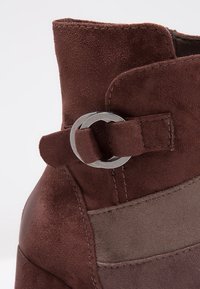 Bottine en daim marron foncé avec une sangle à boucle ronde argentée et un panneau en daim marron clair contrastant.