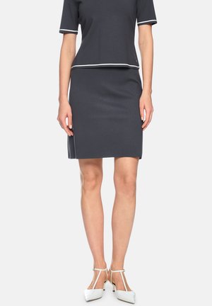 Frau trägt ein dunkelgraues Rock- und Oberteil-Set mit weißen Besätzen, kombiniert mit weißen T-Riemchen-High-Heels, stehend vor einem einfarbigen Hintergrund.