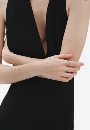 Mujer con un vestido negro sin mangas y escote en V profundo, con los brazos cruzados, luciendo un anillo llamativo plateado y dorado en su mano derecha.