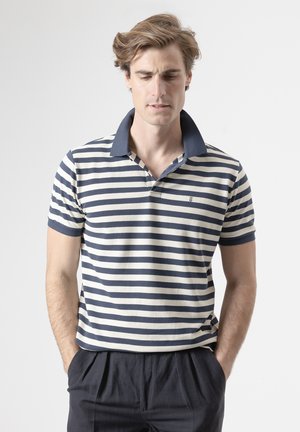 Hombre con camisa polo a rayas azul marino y blanca con cuello azul marino y pantalones oscuros, manos en los bolsillos, mirando hacia abajo.