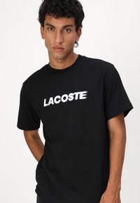 Juodas medvilninis marškinėliai trumpomis rankovėmis, su drąsiu baltu užrašu "LACOSTE" per krūtinę. Paprasta, laisva silueto dizainas.
