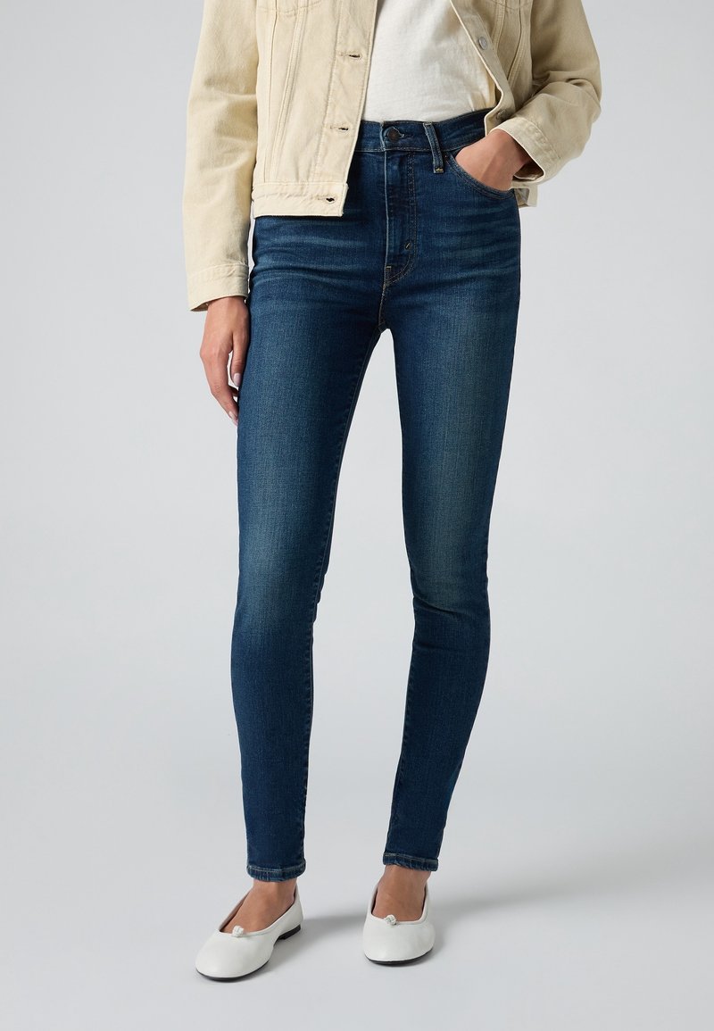 Jean skinny bleu foncé en denim, avec une taille haute, une texture lisse et un effet délavé subtil. Associé à une veste beige clair et des ballerines blanches.