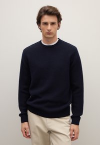 Maglione blu navy lavorato a maglia con un motivo a texture, scollo e polsini a coste. Abbinato a pantaloni beige chiaro.