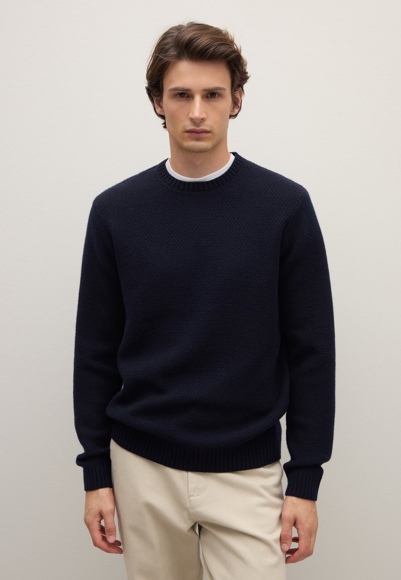 Maglione blu navy lavorato a maglia con un motivo a texture, scollo e polsini a coste. Abbinato a pantaloni beige chiaro.