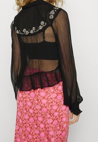 Blusa nera trasparente con polsini arricciati e decorazioni in perline sul colletto, indossata sopra una gonna rosa con stampa floreale.