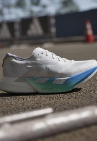 Witte hardloopschoen met blauwe en groene zooldetail, rustend op asfalt nabij een wazige verkeerskegel en barriére in een buitenomgeving.