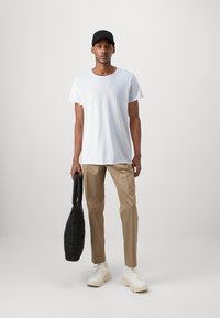 Camiseta blanca de algodón, pantalones beige, gorra negra, sosteniendo un bolso negro acolchado, zapatillas crema con suelas texturizadas, de pie contra un fondo claro.