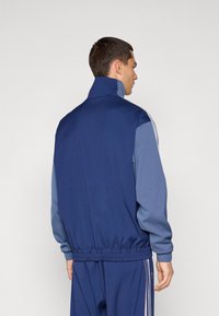 Jachetă de trening navy și albastru deschis, cu guler înalt, talie elastică și dungi laterale. Material moale cu o textură netedă.