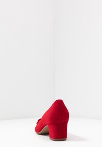 Chaussure à talon en daim rouge avec un bout pointu, une tige texturée et un accent de nœud décoratif sur le côté. Talon carré de taille moyenne pour plus de stabilité.