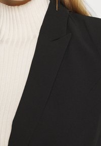 Blazer nero su misura con rever affilati abbinato a un dolcevita a coste color crema. Il tessuto è liscio con un design strutturato e una leggera lucentezza.
