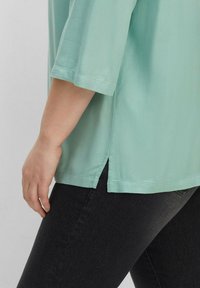 Ljusgrön blus gjord av ett slätt tyg, med trekvartslånga ärmar och sidofickor vid fållen, kombinerad med mörka jeans.