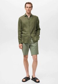 Light green linen long-sleeve shirt, relaxed fit, button-down front. Paired with light green shorts and black sandals. Smooth texture.