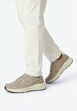Voeten in beige en crèmekleurige sportieve sneakers met dikke zolen, gecombineerd met witte broeken tegen een effen lichte achtergrond.