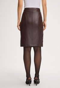 Bruine leren kokerrok met een rechte snit, zichtbare stiksels en een hoge tailleband, gecombineerd met zwarte doorschijnende panty’s en hakken.