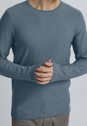 Homme portant une chemise bleue à longues manches avec des ouvertures pour les pouces, les mains jointes devant, sur un fond clair et uni.