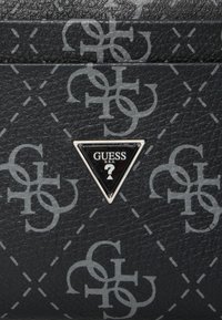 Material negro texturizado con un patrón repetido de logotipos grises. Presenta una placa triangular plateada con "GUESS" en el centro.