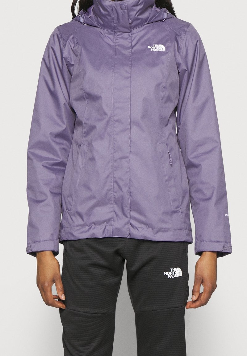 Personne portant une veste imperméable North Face violette et un pantalon North Face noir, debout les mains détendues sur les côtés.